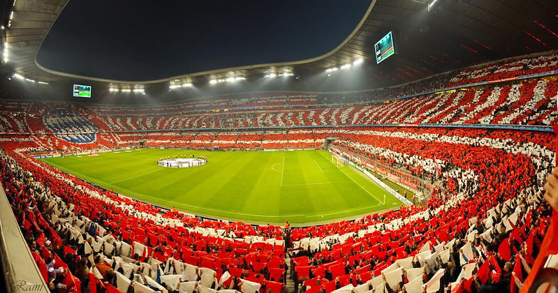 a full stadium with a soccer pitch with the champions league badge in the center circle, fans holding up red and white shirts to make red and white lines and the bayern munich badge